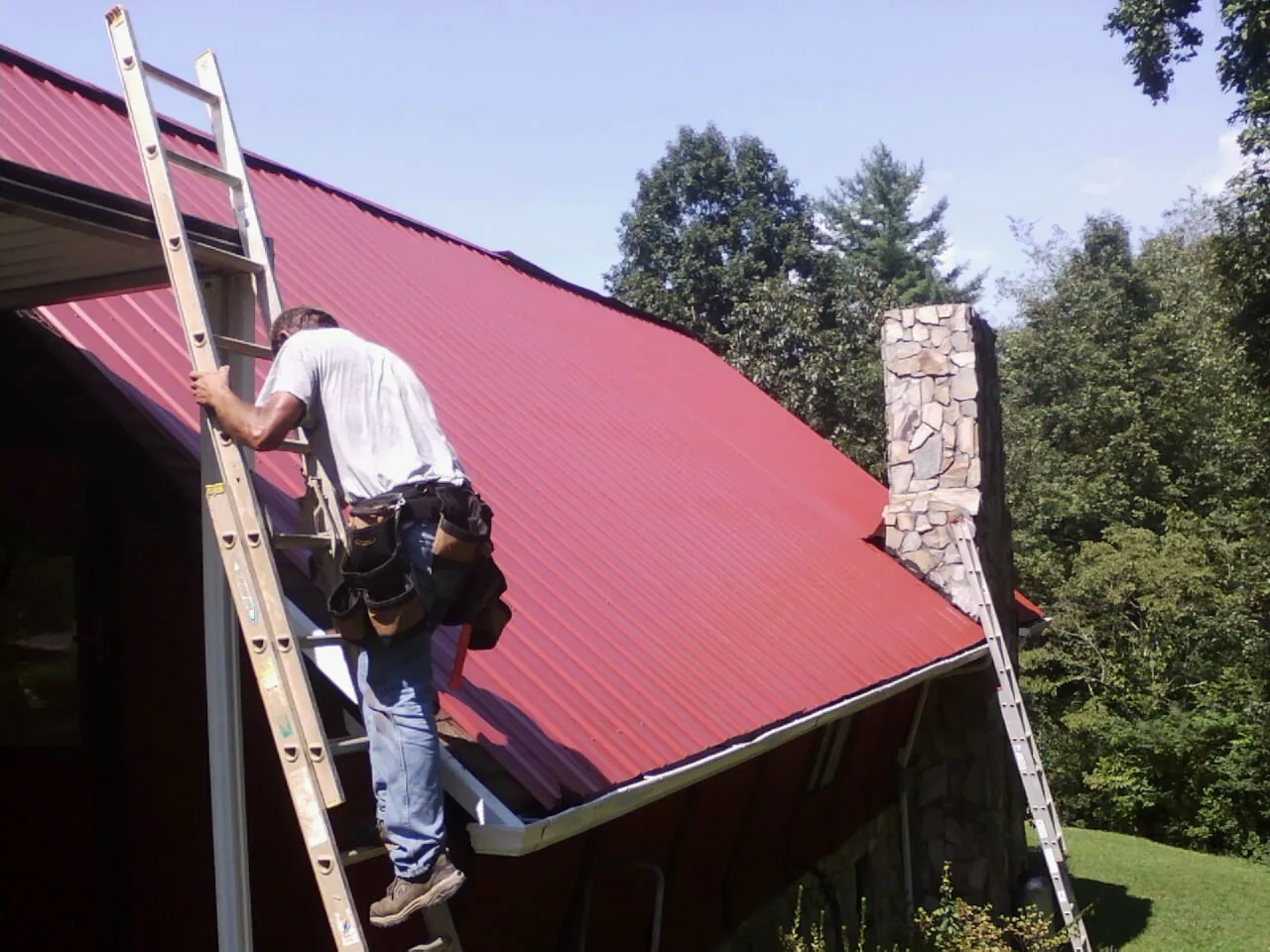 Roofer on a ladder beside a red metal roof and stone chimney — Soffit & Fascia Repair by Roofing Brothers in Groveland